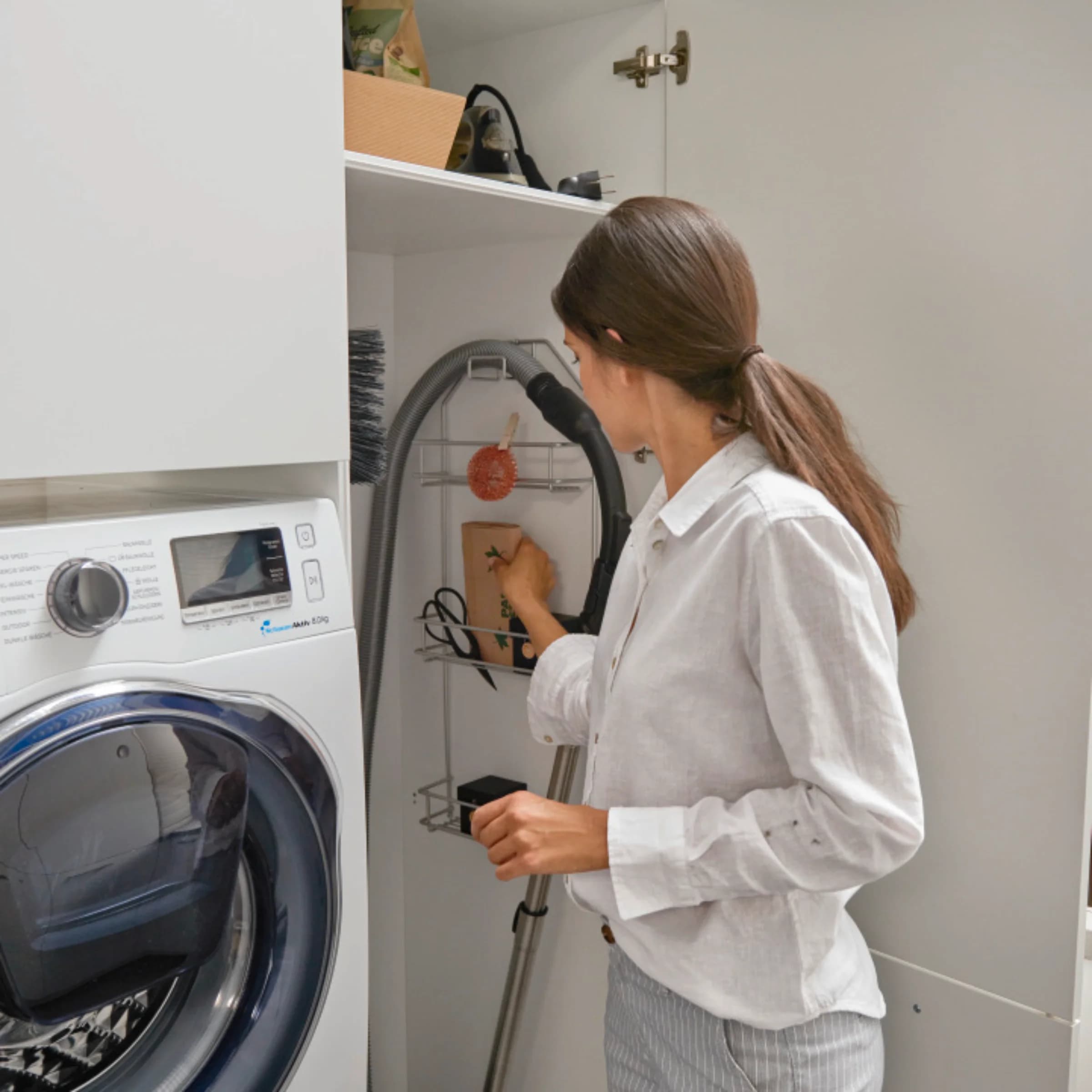 A woman organizes a utility room with a vacuum, washing machine, and shelves. Nolte utility rooms.